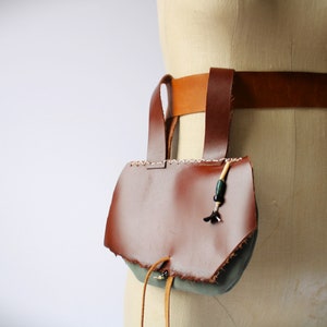 May include: A brown leather pouch with a flap and a drawstring closure. The pouch is attached to a brown leather belt with a buckle. The pouch has a green fabric lining and a beaded tassel.