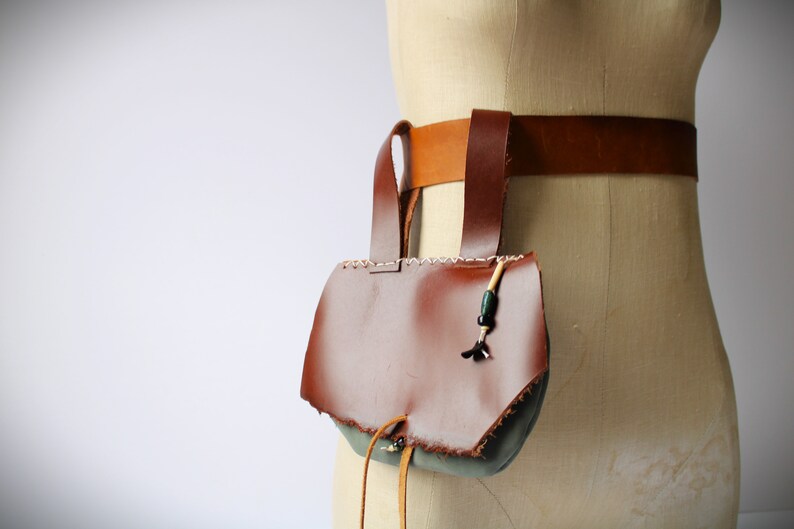 May include: A brown leather pouch with a flap and a drawstring closure. The pouch is attached to a brown leather belt with a buckle. The pouch has a green fabric lining and a beaded tassel.