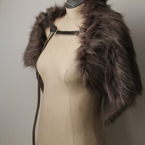 May include: A gray faux fur shrug with a leather strap. The shrug is displayed on a mannequin against a white background.