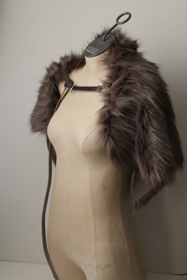 May include: A gray faux fur shrug with a leather strap. The shrug is displayed on a mannequin against a white background.