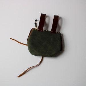 May include: A small, green leather bag with brown leather straps and a drawstring closure. The bag is made of suede and has a rustic, handmade look.