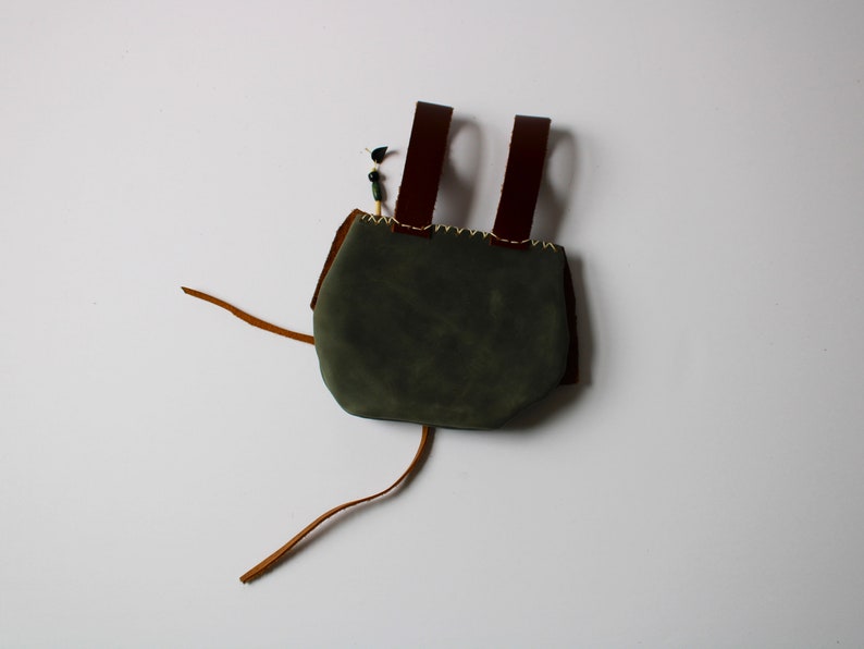 May include: A small, green leather bag with brown leather straps and a drawstring closure. The bag is made of suede and has a rustic, handmade look.