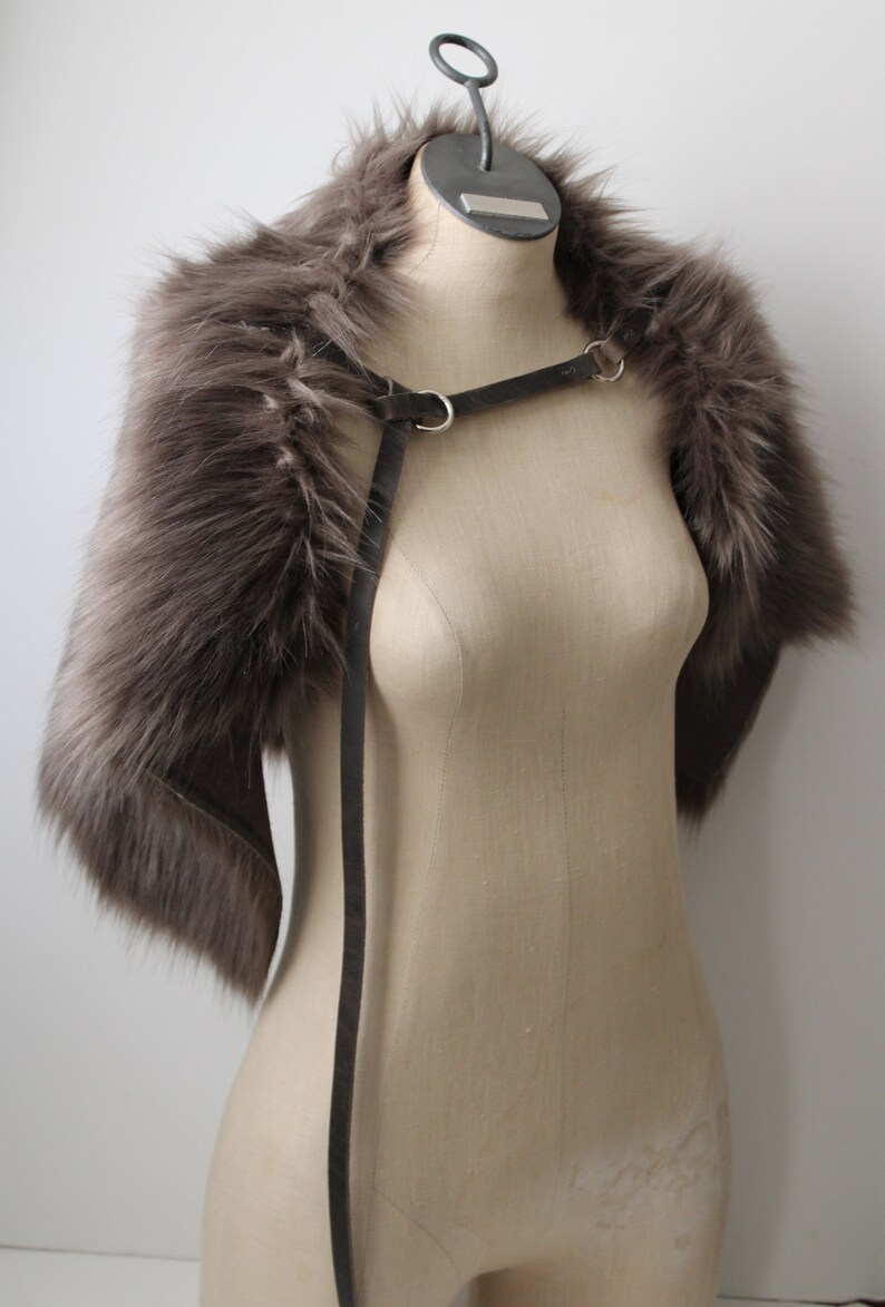 May include: A gray faux fur stole with a black leather strap and silver buckles. The stole is displayed on a mannequin.