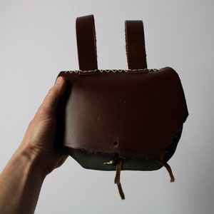 May include: A brown leather pouch with two straps and a drawstring closure. The pouch is held in a hand.