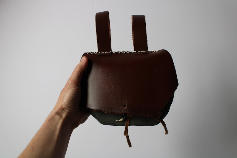 May include: A brown leather pouch with two straps and a drawstring closure. The pouch is held in a hand.