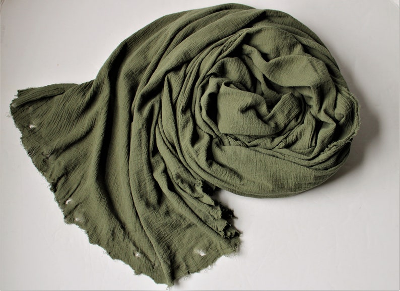 the drab green cotton shawl is spun in a spiral on a white background to showcase the deliberately distressed edges
