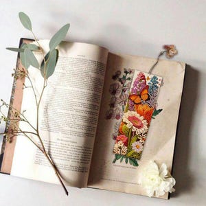 May include: A colorful bookmark with a floral design and butterflies. The bookmark is made of fabric and has a tassel on the top. It is sitting on an open book.