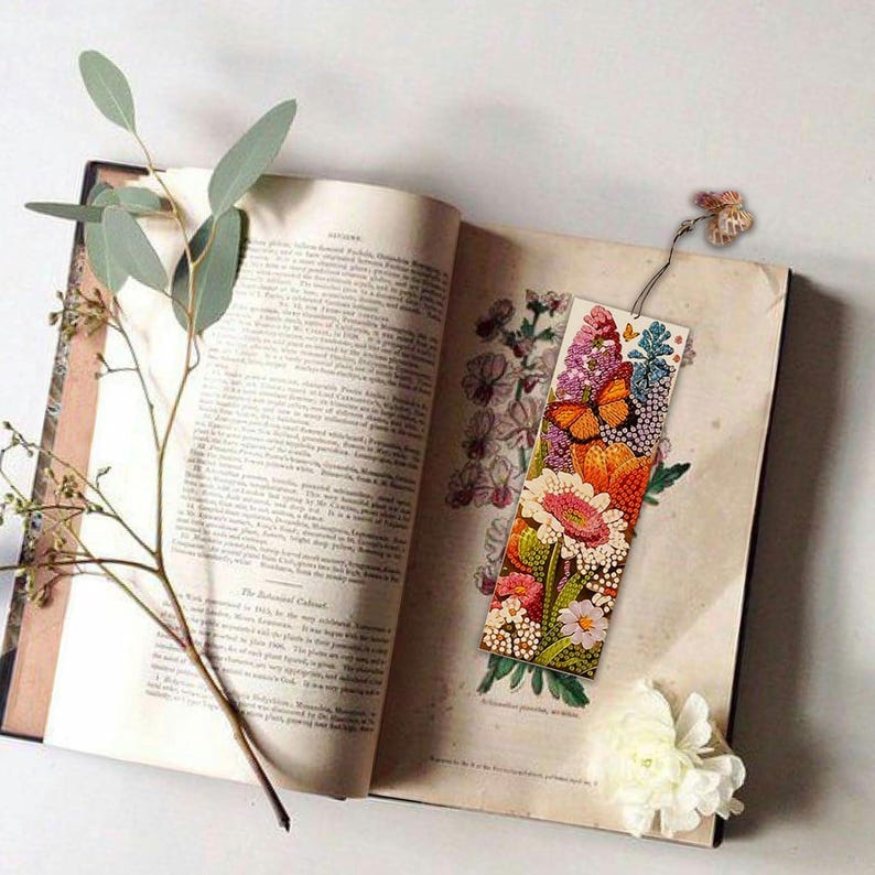 May include: A colorful bookmark with a floral design and butterflies. The bookmark is made of fabric and has a tassel on the top. It is sitting on an open book.