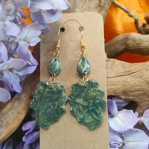 Op de afbeelding: Handgemaakte groene en gouden oorbellen met een bloemmotief. De oorbellen worden gepresenteerd op een bruine oorbellenkaart met goudkleurige oorhaken. De achtergrond omvat paarse bloemen, drijfhout en een oranje achtergrond.