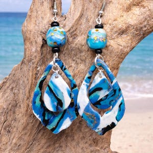 Op de afbeelding: Handgemaakte oorbellen met een strandthema. De oorbellen hebben een blauw, wit en zwart gemarmerd ontwerp met gouden accenten. De bovenste kraal is een blauwe en gouden bol. De oorbellen zijn opgehangen aan zilverkleurige haken.