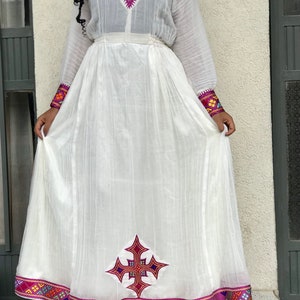 May include: A white traditional Ethiopian dress with colorful embroidery around the neckline and hem. The dress features a cross design on the front and a purple waistband.