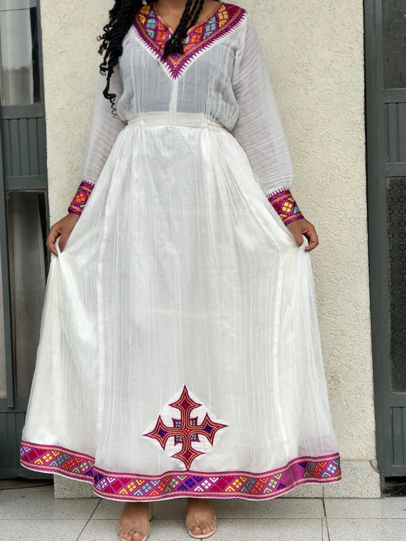 May include: A white traditional Ethiopian dress with colorful embroidery around the neckline and hem. The dress features a cross design on the front and a purple waistband.