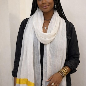 May include: A white scarf with a gold border is draped around the neck. The scarf is paired with a black jacket and gold bracelets. The scarf is a fashion accessory.
