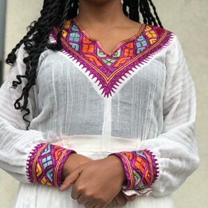 May include: White long-sleeved dress with colorful embroidered details around the neckline and cuffs. The embroidery features geometric patterns in shades of pink, blue, green, orange, and yellow.