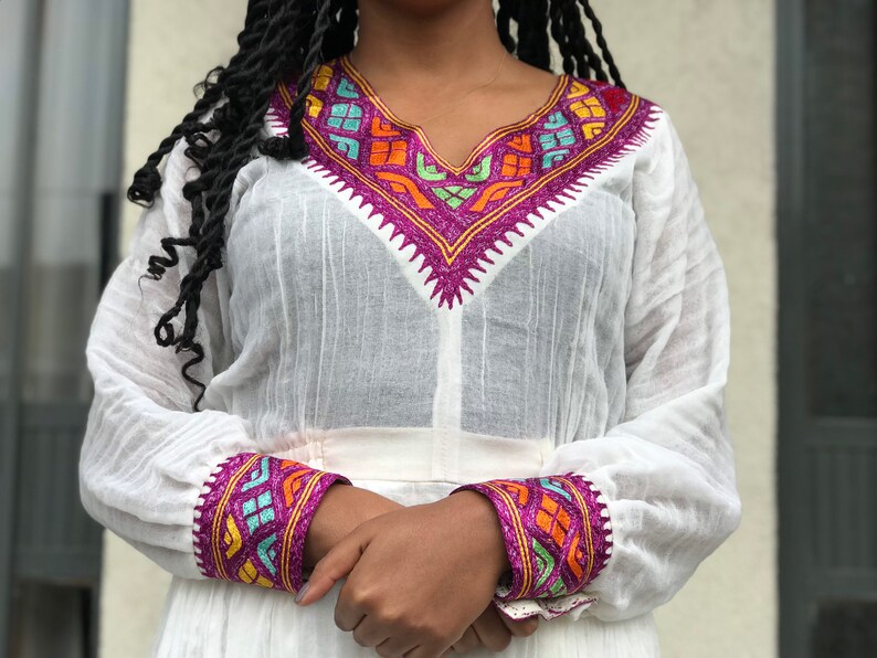 May include: White long-sleeved dress with colorful embroidered details around the neckline and cuffs. The embroidery features geometric patterns in shades of pink, blue, green, orange, and yellow.