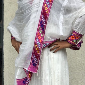 May include: A white traditional Ethiopian dress with a pink and multi-colored embroidered trim. The dress is made of a lightweight fabric and has a flowing silhouette.