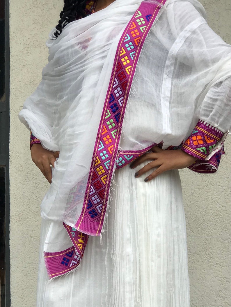 May include: A white traditional Ethiopian dress with a pink and multi-colored embroidered trim. The dress is made of a lightweight fabric and has a flowing silhouette.