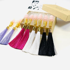 May include: Tassel earrings in shades of purple, fuchsia, white, and black. Each earring has a gold-coloured decorative cap and a pink, cone-shaped ear plug. The earrings are presented in a small, brown box.