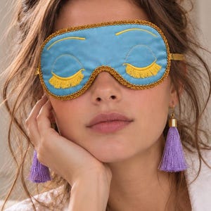 Puede incluir: Un antifaz para dormir de color azul claro con ojos cerrados bordados, pestañas largas amarillas y un borde trenzado dorado. El antifaz se sujeta con una fina cinta dorada y se combina con pendientes de borlas moradas con herrajes dorados.