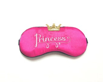 Può includere: Maschera per dormire in raso rosa con una corona dorata e la parola "Princess" ricamata in bianco.