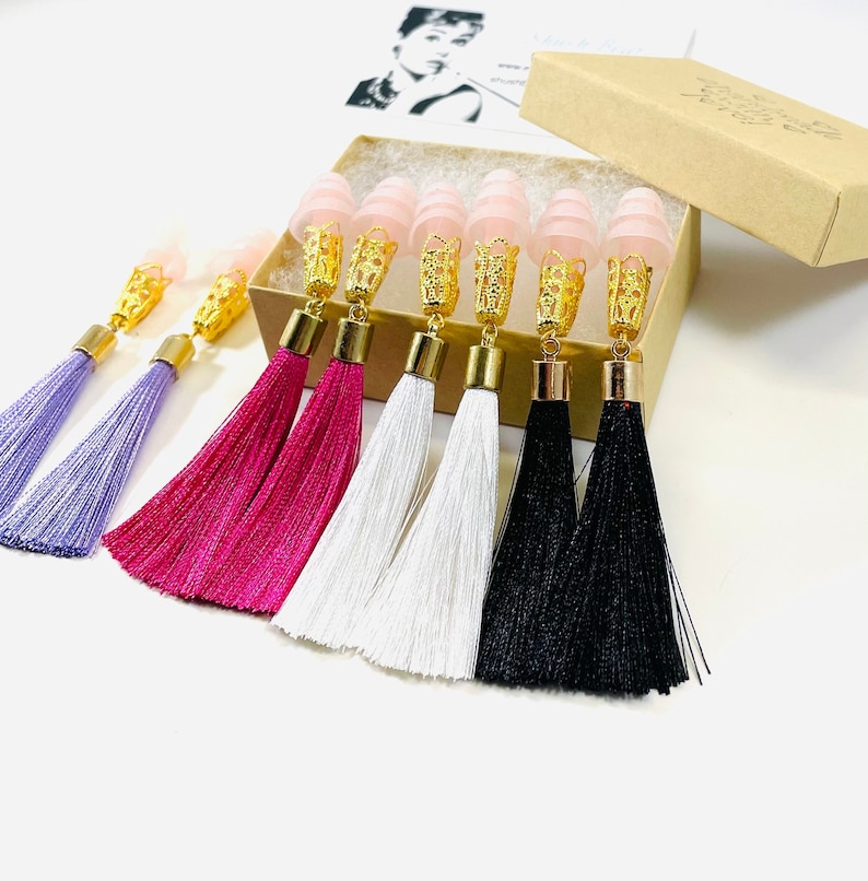 May include: Tassel earrings in shades of purple, fuchsia, white, and black. Each earring has a gold-coloured decorative cap and a pink, cone-shaped ear plug. The earrings are presented in a small, brown box.