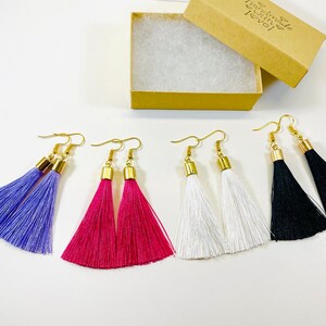 May include: Tassel earrings in shades of purple, fuchsia, white, and black, each with a gold-tone cap and hook. The earrings are displayed alongside an open cardboard box, showcasing the handmade jewellery.