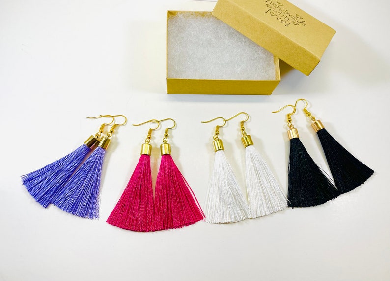 May include: Tassel earrings in shades of purple, fuchsia, white, and black, each with a gold-tone cap and hook. The earrings are displayed alongside an open cardboard box, showcasing the handmade jewellery.