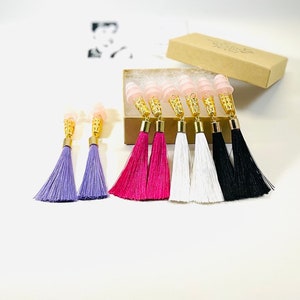May include: Earplugs with gold filigree and tassel earrings in purple, fuchsia, white, and black. The earplugs are pink and are in a small cardboard box. The earrings are displayed on a white surface.