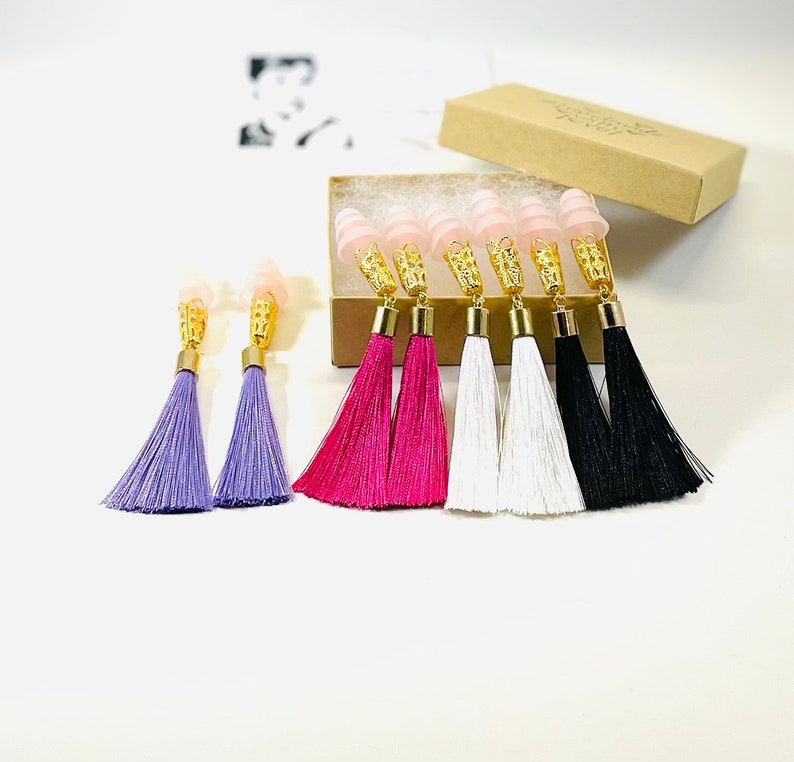 May include: Earplugs with gold filigree and tassel earrings in purple, fuchsia, white, and black. The earplugs are pink and are in a small cardboard box. The earrings are displayed on a white surface.