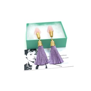 Könnte beinhalten: Ein Paar violette Quastenohrringe mit rosa und goldenen Akzenten. Die Ohrringe werden in einer hellgrünen Schachtel mit einer Visitenkarte mit der Aufschrift "Shushbear" präsentiert.