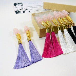 May include: Ear plugs with pink, gold, and tassel details. The ear plugs have a light pink, cone-shaped top, gold filigree, and a gold base. The tassels are in purple, pink, white, and black. A business card with an Audrey Hepburn image is in the background.