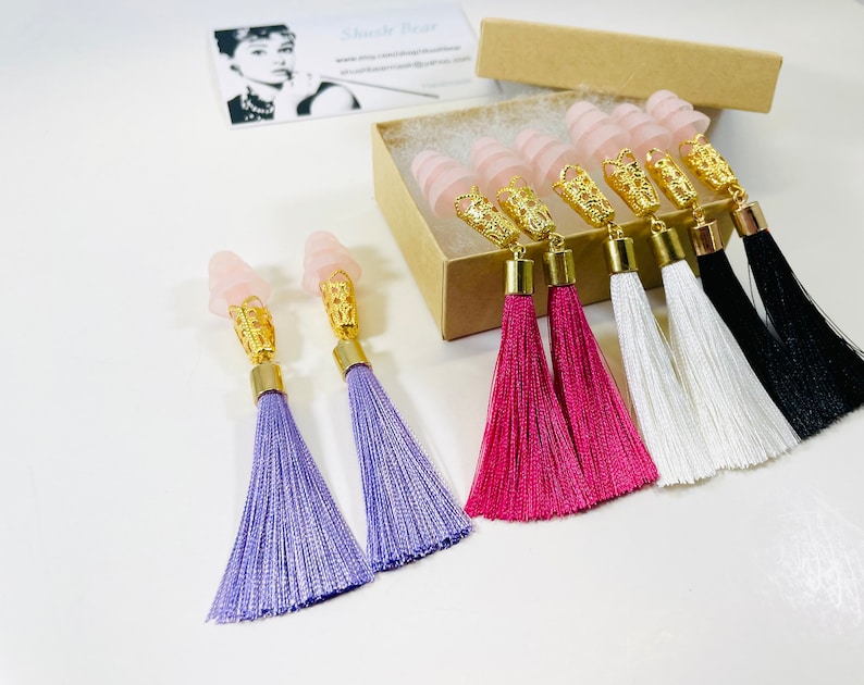 May include: Ear plugs with pink, gold, and tassel details. The ear plugs have a light pink, cone-shaped top, gold filigree, and a gold base. The tassels are in purple, pink, white, and black. A business card with an Audrey Hepburn image is in the background.