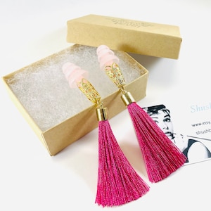 May include: A pair of pink tassel earrings with gold-coloured filigree accents and light pink earplugs. The earrings are displayed in a small, open cardboard box with white padding.
