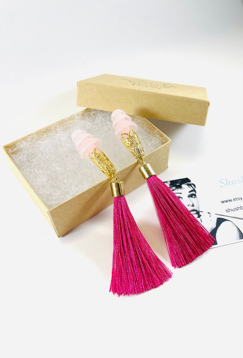 May include: A pair of pink tassel earrings with gold-coloured filigree accents and light pink earplugs. The earrings are displayed in a small, open cardboard box with white padding.