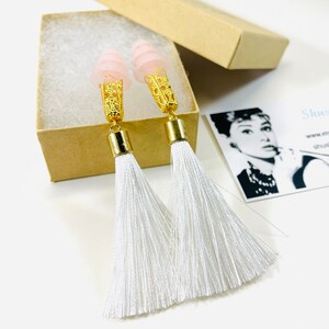 May include: A pair of long tassel earrings with pink tops and gold-coloured filigree accents. The white tassels hang from gold-coloured cylinders. The earrings are displayed in an open, small, brown cardboard box.
