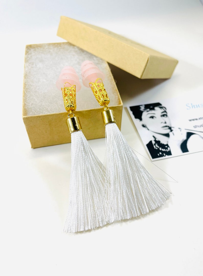 May include: A pair of long tassel earrings with pink tops and gold-coloured filigree accents. The white tassels hang from gold-coloured cylinders. The earrings are displayed in an open, small, brown cardboard box.