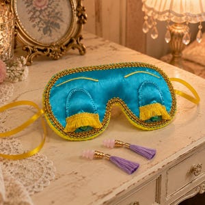 Könnte beinhalten: Eine leuchtend blaue Schlafmaske aus Satin mit goldener Flechtborte, gelben Fransen und gelben Bandbindern, begleitet von zwei Ohrstöpseln mit hellvioletten Quasten und rosa Silikonspitzen zur Licht- und Schallabschirmung.