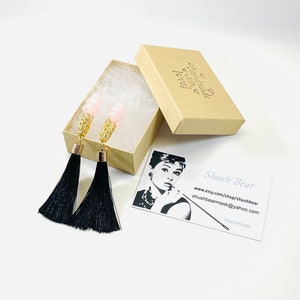 May include: Dangle earrings featuring pink beads, gold filigree, and long black tassels, displayed in an open cardboard box with tissue paper. A business card with an Audrey Hepburn image is also present.