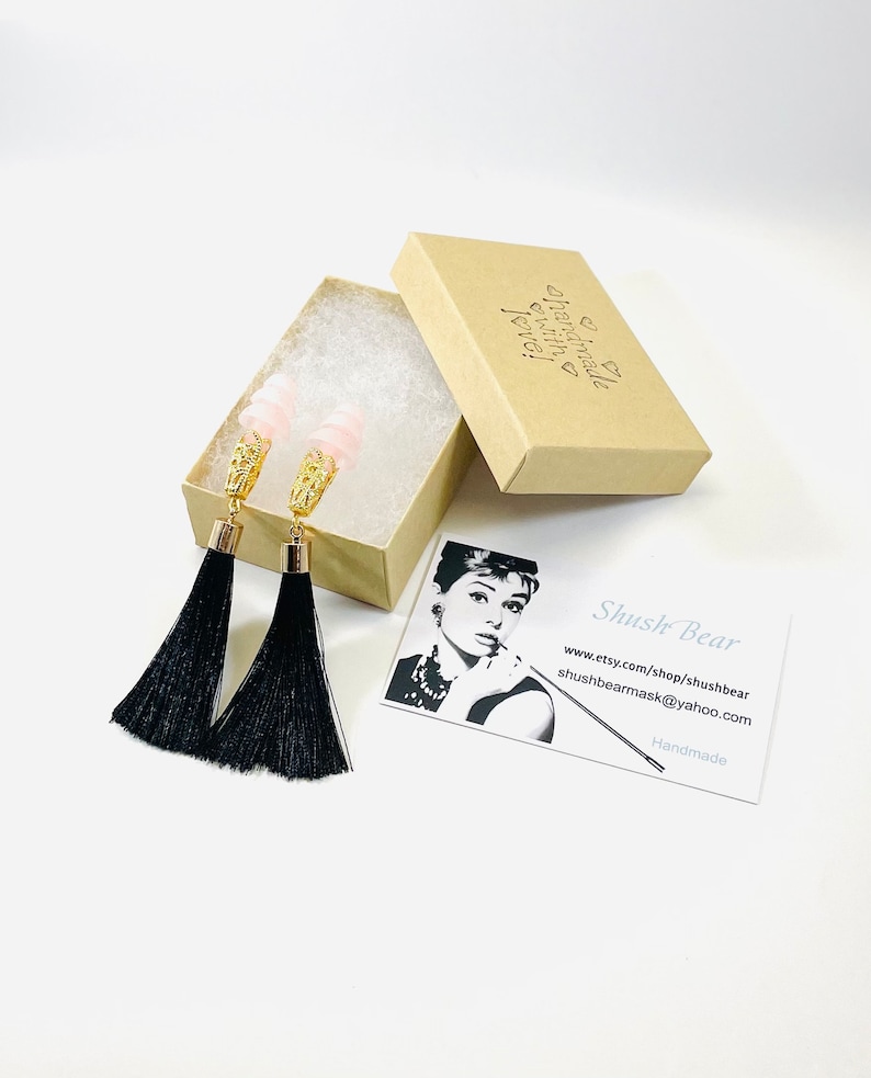 May include: Dangle earrings featuring pink beads, gold filigree, and long black tassels, displayed in an open cardboard box with tissue paper. A business card with an Audrey Hepburn image is also present.