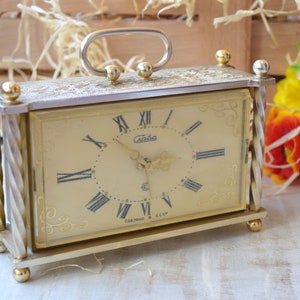 May include: A gold-toned alarm clock with a white face and black Roman numeral hour markers. The clock has a silver frame with a decorative design and a handle on the top. The clock is made in the USSR.