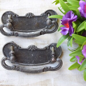 Puede incluir: Dos tiradores de cajón de plata antigua con detalles ornamentados y un patrón de panal. Los tiradores tienen una forma curva y están diseñados para su uso en armarios o muebles.