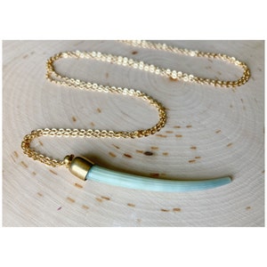 Puede incluir: Un collar de cadena dorada con un colgante. El colgante es un diseño en forma de cuerno azul claro con detalles metálicos dorados. El collar se muestra sobre una superficie de madera marrón claro.