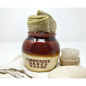 May include: A brown and beige ceramic bean pot with the words "Tennessee Baked Beans" printed on the front. The pot contains folded green and brown woven washcloths. A small wooden brush and a bar of soap are next to the pot.