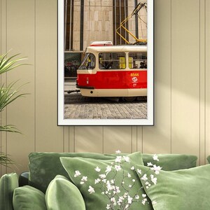 May include: Framed print of a red and cream tram, number 8544, against a stone building and cobbled street. The black-framed image hangs above a green velvet sofa. The tram's yellow details contrast with the red and cream.
