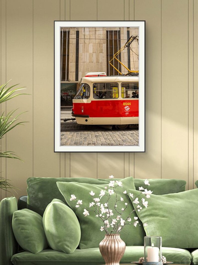 May include: Framed print of a red and cream tram, number 8544, against a stone building and cobbled street. The black-framed image hangs above a green velvet sofa. The tram's yellow details contrast with the red and cream.