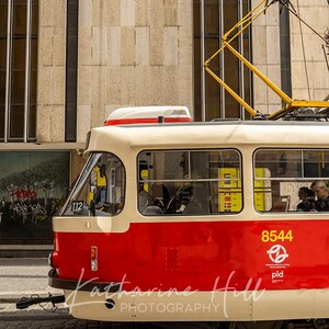 May include: A red and cream tram car, number 8544, on a cobbled street. The tram has a yellow overhead electrical system. The building behind is beige. The tram is a mode of public transport.