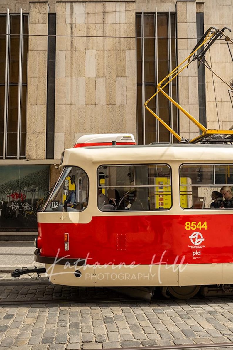 May include: A red and cream tram car, number 8544, on a cobbled street. The tram has a yellow overhead electrical system. The building behind is beige. The tram is a mode of public transport.