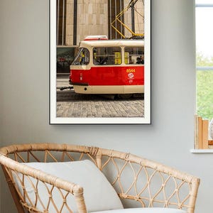 May include: Framed photograph of a red and cream tram with the number 8544 on the side. The tram is on a cobbled street in front of a beige building. The tram's yellow electrical lines are visible. The photograph is hanging above a wicker chair.