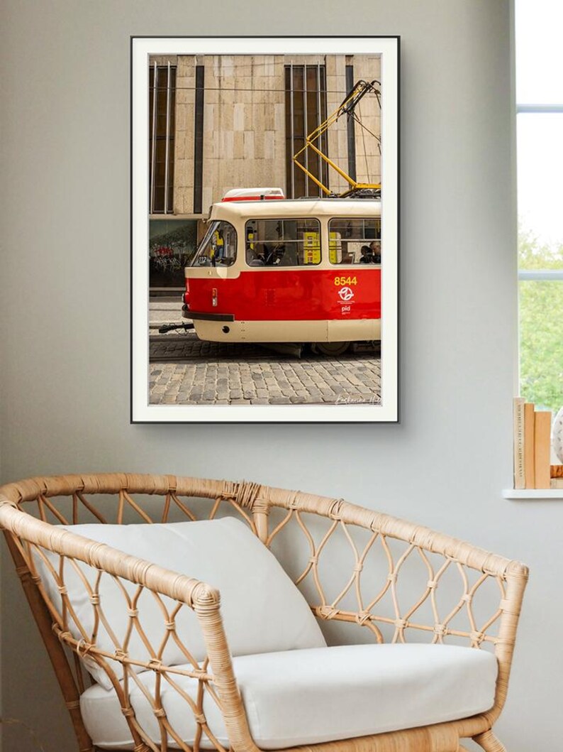 May include: Framed photograph of a red and cream tram with the number 8544 on the side. The tram is on a cobbled street in front of a beige building. The tram's yellow electrical lines are visible. The photograph is hanging above a wicker chair.