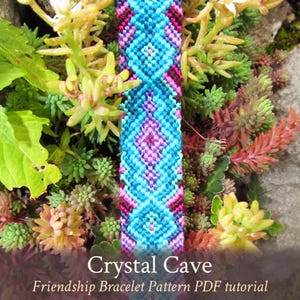 Könnte beinhalten: Ein farbenfrohes Freundschaftsarmband mit dem Muster "Crystal Cave". Das Armband ist aus blauer, rosa, violetter und brauner Stickgarn hergestellt. Das Armband liegt auf einem Bett aus grünen und roten Sukkulenten.